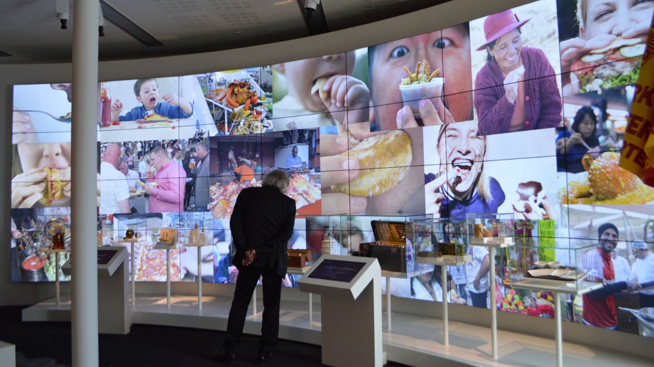 A man stands in front of a large screen which shows various images of people interacting with food.
