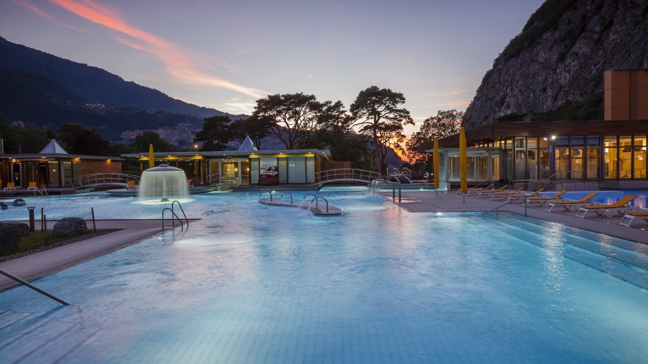 Piscine extérieure d'un centre thermal en soirée, avec une grande fontaine éclairée au centre, plusieurs ponts arqués enjambant les bassins et des chaises longues disposées le long de la piscine. Le site est entouré de montagnes et d’arbres avec un ciel coloré par le coucher du soleil.