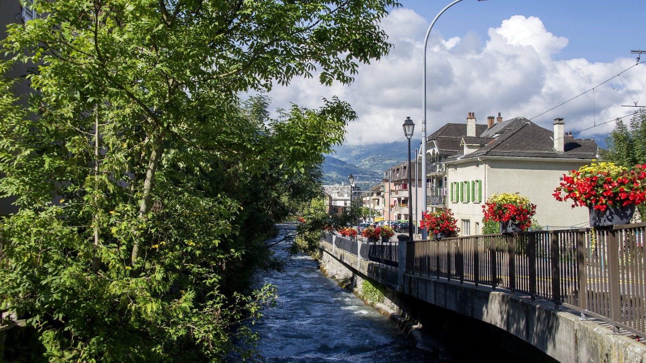 À gauche de l’image, des arbres donnent leur ombre à un canal. À droite, une route bordée de maisons aux murs blancs et volets verts. En arrière-plan, on devine des montagnes surplombées de quelques nuages et un ciel bleu.