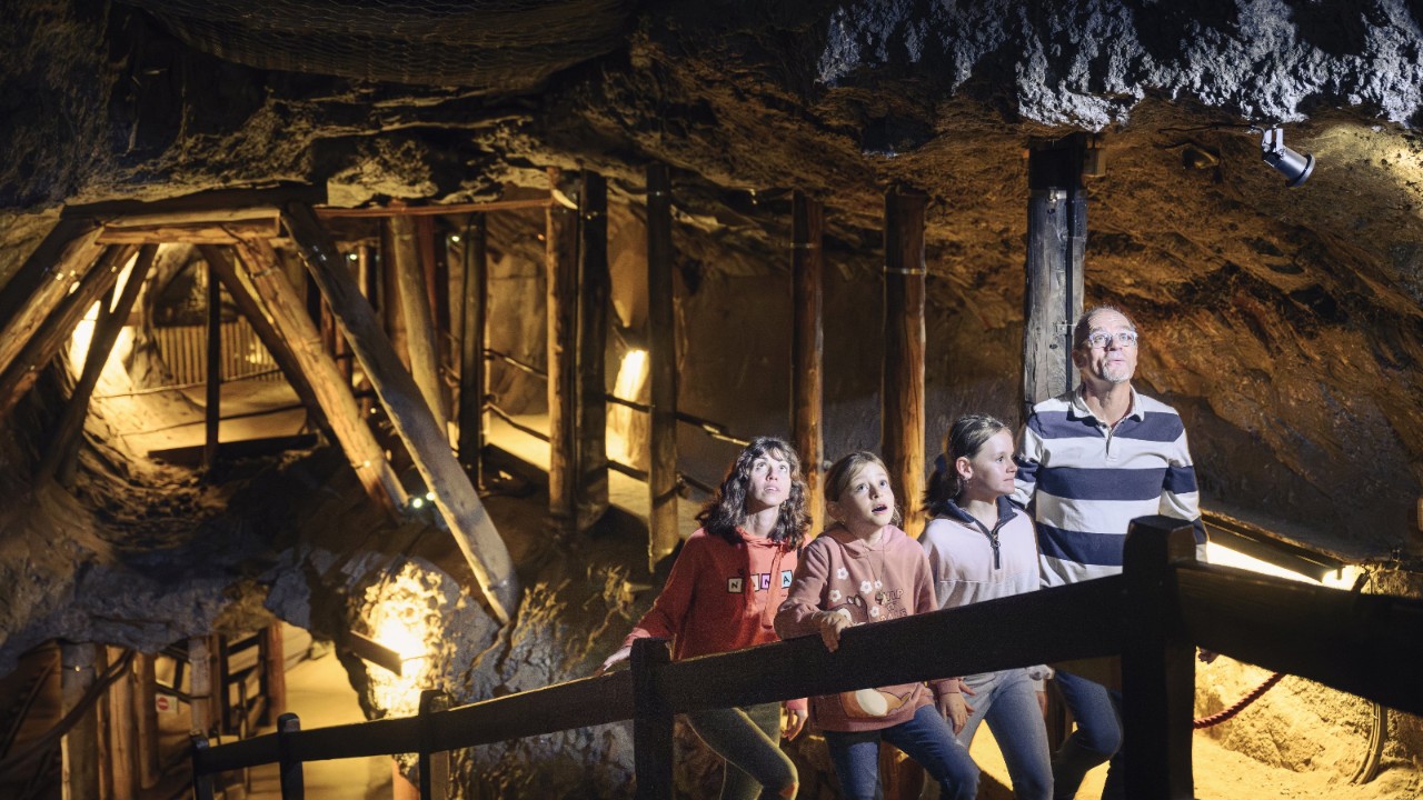 Une famille de quatre personnes explore une galerie souterraine éclairée. Autour d’elles, des parois rocheuses et des poutres en bois qui soutiennent le plafond de la mine. Les enfants et les adultes regardent vers le haut, fascinés par ce qu’ils découvrent.