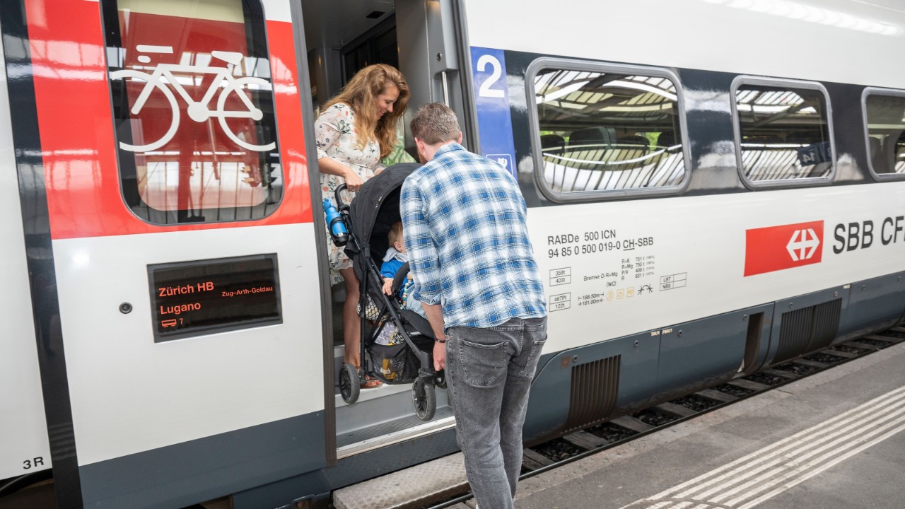 Drehen Sie den Kinderwagen mit Lenker Richtung Zug. Eine Person geht zuerst hinein, nimmt oben den Lenker in die Hand und die 2. Person bleibt unten stehen und hält eine Querstange bei den Rädern. Heben Sie dann gemeinsam den Kinderwagen in den Zug.