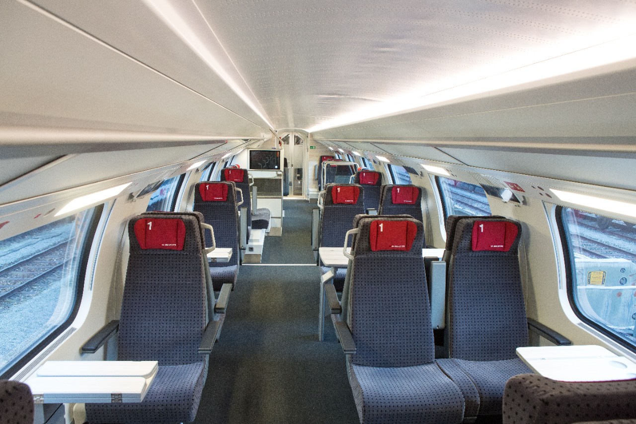 Allestimento interno di 1ª classe – A destra del corridoio centrale si trovano i compartimenti da quattro posti e sulla sinistra quelli da due. I sedili sono di colore grigio scuro con piccoli cuscini rossi sui poggiatesta.