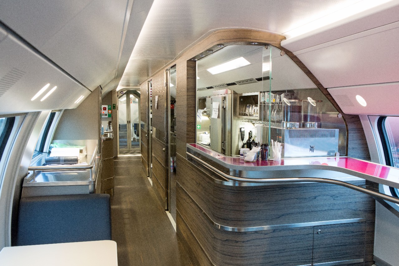 Carrozza ristorante sull’IC 200 – Sulla destra dell’immagine si scorgono la cucina e il bancone. Il pavimento e la cucina sono realizzati in legno, la superficie d’appoggio del bancone è rosa fucsia. Sulla sinistra del corridoio si notano una superficie d