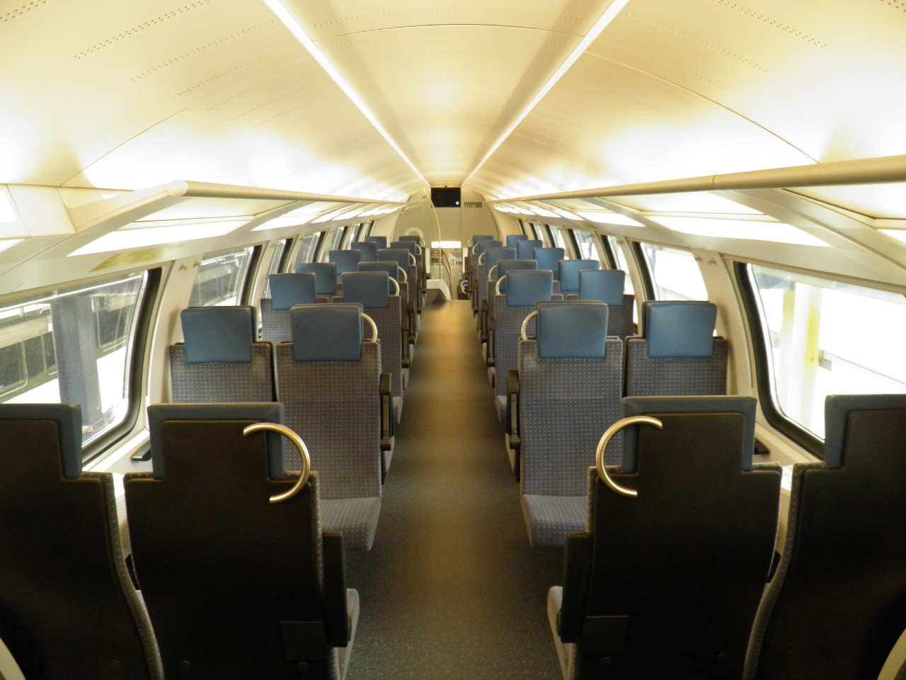 IR-Dosto: Innenansicht der zweiten Klasse. Im Fokus die vierer Abteile der zweiten Klasse mit Blauer Kopflehne und Blau gepunktetem Sitzstoff. Am ende des Wagens, obehalb dem Abstieg in die untere Ebene, befindet sich ein Bildschirm für Kundeninformationen.