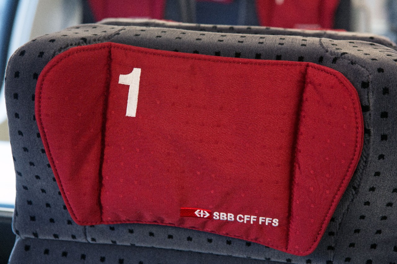 Allestimento interno di 1ª classe – A sinistra del corridoio centrale si trovano i compartimenti da quattro posti e a sinistra quelli da due. I sedili sono di colore grigio scuro con piccoli cuscini rossi sui poggiatesta.